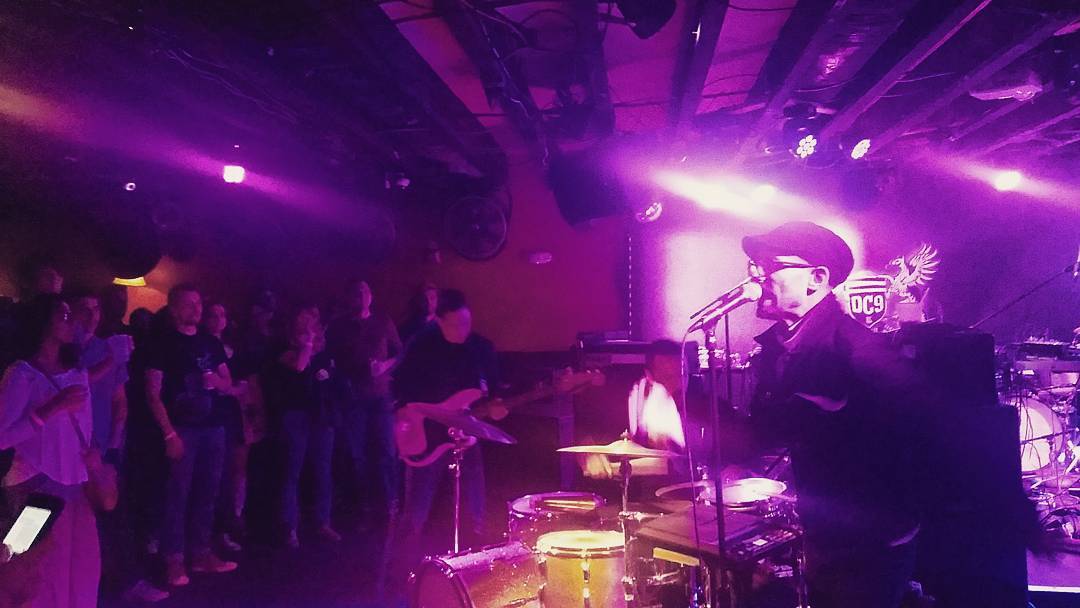 On the right a singer uses his microphone, standing in front of a drumset and guitarist and the DC9 sign. On the left stands the crowd, all same level on the floor.